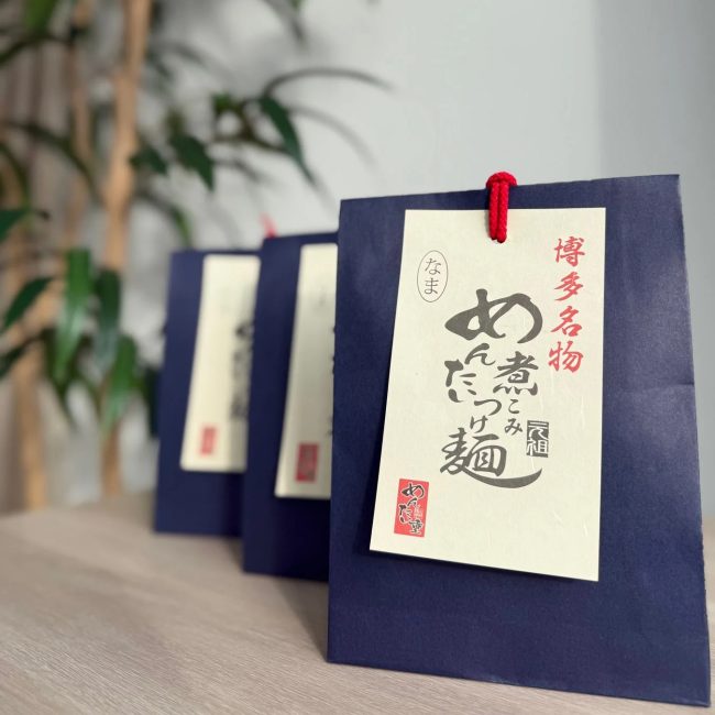 紺色のパッケージに「博多名物 めんたいつけ麺」と書かれた札がかけられた贈答品が複数並んでいます。お土産や贈り物に最適な食品であることが伺えます。